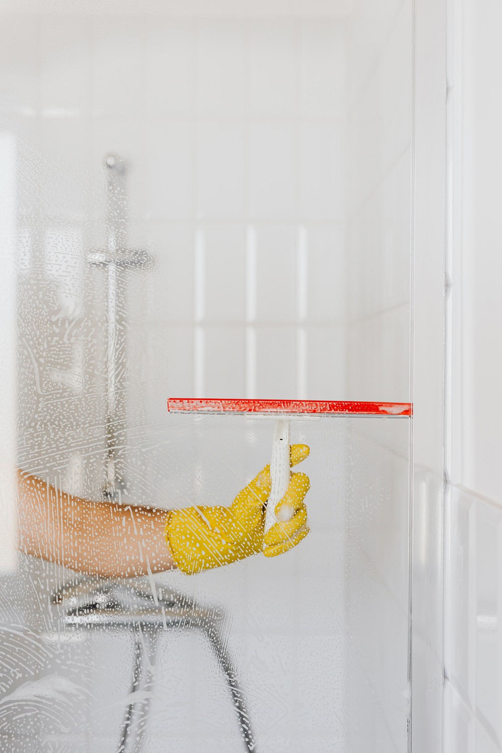 L’aceto bianco aiuta una donna a pulire il vetro del box doccia dal calcare. La donna asciuga le gocce d’acqua con un tergivetri mentre indossa i guanti.